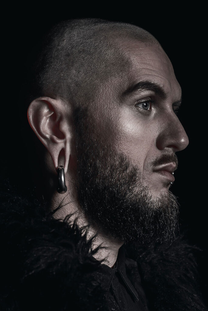 Porträt eines Mannes mit Bart und Tunnel-Piercing im Ohr, dargestellt in einem dunklen, stimmungsvollen Bild.